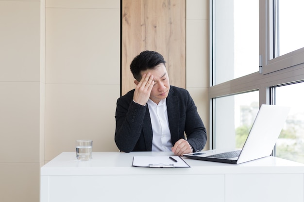 Un jeune employé de bureau asiatique souffrant de maux de tête au travail au bureau boit une pilule médicamenteuse avec de l'eau. Un homme malade en costume devant l'ordinateur à l'intérieur avec une douleur intense utilise le médicament sur le lieu de travail