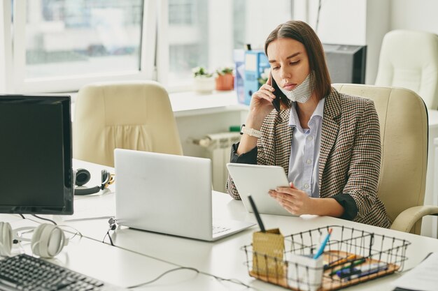 Jeune employé de bureau jolie brune avec masque de protection sur le menton à la recherche de texte sur l'écran du pavé tactile et au client de téléphoner tout en travaillant