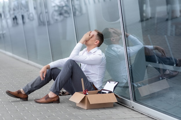 Un jeune employé de bureau licencié est assis sous son bureau. À côté de lui se trouve sa papeterie. L'homme est stressé.