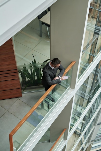 Jeune employé masculin en costume debout près de la balustrade dans le centre de bureau et envoyer des SMS