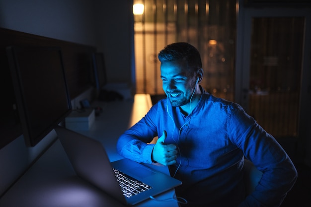 Jeune employé de race blanche ayant un appel vidéo avec le patron et montrant le pouce vers le haut tout en étant assis dans le bureau tard dans la nuit.