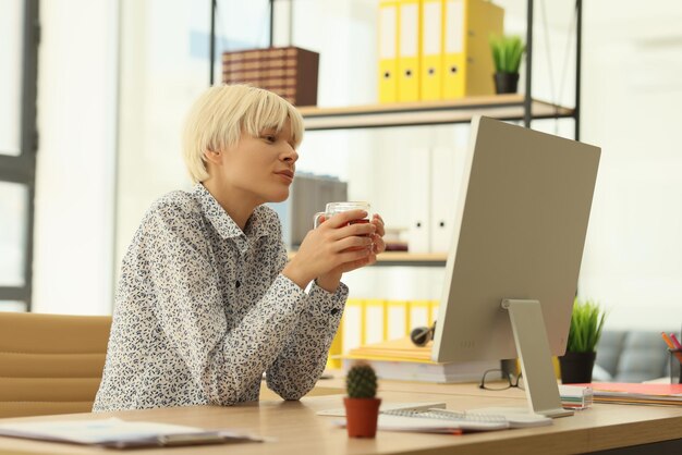Une jeune employée de bureau boit du thé chaud assis à table avec un ordinateur et un employé de cactus