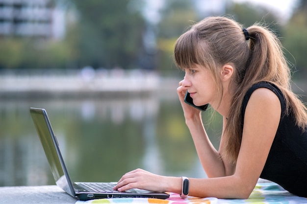 Jeune employée de bureau travaillant derrière un ordinateur portable tout en parlant au téléphone portable allongée dans un parc d'été à l'extérieur. Concept de travail à distance.