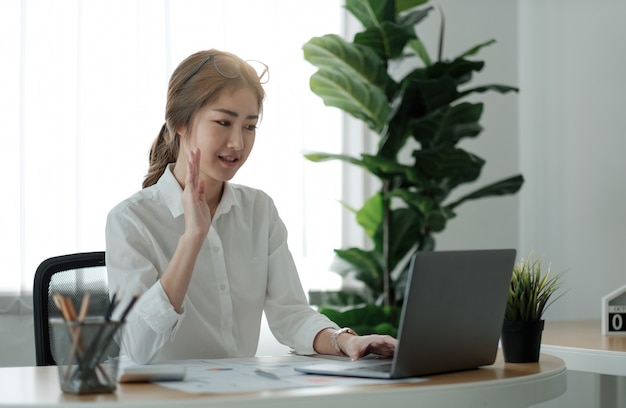 Une jeune employée souriante à la maison fait un signe de la main lors d'un appel vidéo sur un ordinateur portable avec divers collègues. Une travailleuse asiatique a une conférence par webcam ou une réunion d'équipe Web numérique ou un briefing avec des collègues.