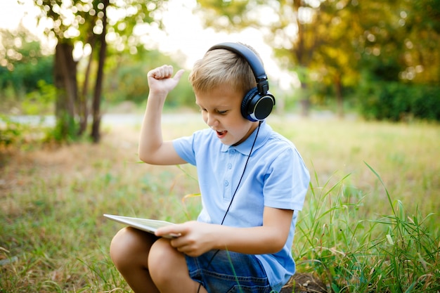 Jeune enfant assis dans la forêt portant des écouteurs et jouant à un jeu sur tablet pc.