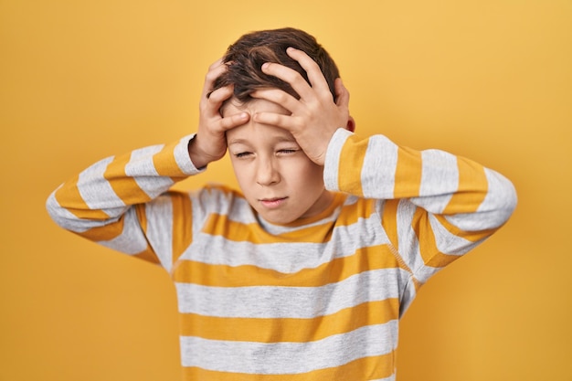 Jeune enfant caucasien debout sur fond jaune souffrant de maux de tête désespérés et stressés à cause de la douleur et de la migraine. mains sur la tête.