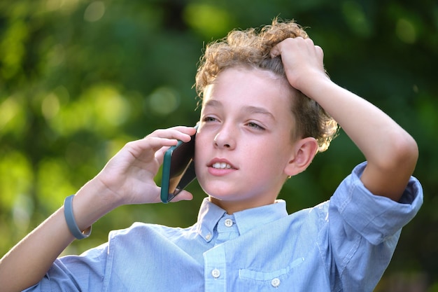 Jeune enfant garçon perplexe parlant sur téléphone portable à l'extérieur dans le parc d'été