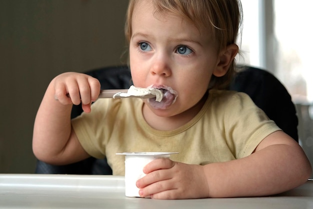 Jeune enfant mangeant un mélange d'aliments en purée assis dans une chaise haute bébé sevrant petite fille apprenant à manger du yaourt se nourrissant petite main avec une cuillère petit-déjeuner avec un produit laitier enfant premier aliment solide