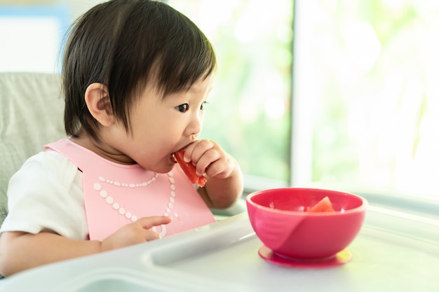 Jeune enfant mignon sur une chaise haute pour bébé, tenant un melon d'eau avec un visage souriant à la maison, profitez d'un repas avec bonheur.