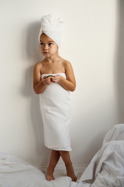 Un jeune enfant de race blanche aux cheveux enveloppés dans des serviettes de bain blanches appliquant des morceaux de concombre à ses yeux sur un fond blanc