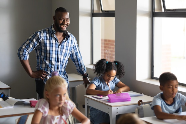 Jeune enseignant afro-américain souriant et élèves multiraciaux du primaire au bureau dans la salle de classe