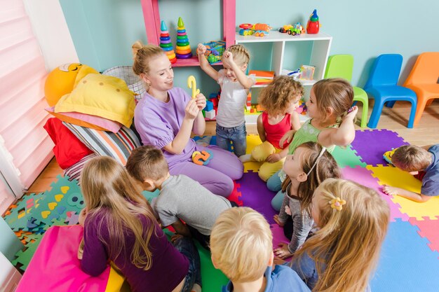 Jeune enseignant engagé assis avec un groupe d'enfants d'âge préscolaire sur un sol pendant la leçon Le processus d'apprentissage peut être amusant Salle avec des tapis colorés sur un sol et des meubles multicolores