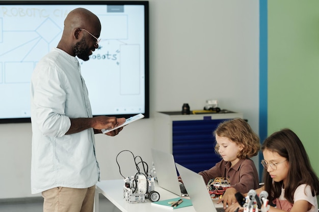 Jeune enseignant avec tablette debout devant des écoliers avec des ordinateurs portables