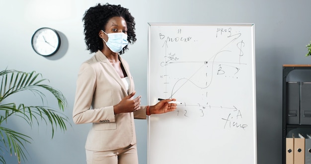 Jeune enseignante afro-américaine en masque médical debout à bord en classe et en disant les lois de la physique ou de la géométrie à la classe. Concept de pandémie. École pendant le coronavirus. Lection éducative.