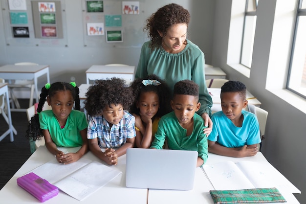 Une jeune enseignante blanche debout à côté d'étudiants élémentaires afro-américains utilisant un ordinateur portable à leur bureau.