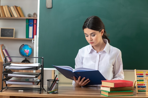 Jeune enseignante confiante lire le dossier assis à table avec des outils scolaires en classe