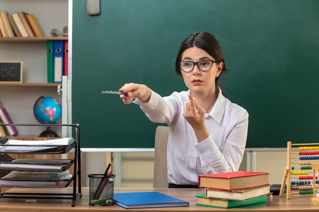 Une jeune enseignante impressionnée portant des lunettes pointe sur le côté avec un bâton de pointeur assis à table avec des outils scolaires en classe