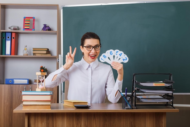 Jeune enseignante joyeuse portant des lunettes tenant des plaques d'immatriculation expliquant la leçon montrant vsign tirant la langue assis au bureau de l'école devant le tableau noir dans la salle de classe