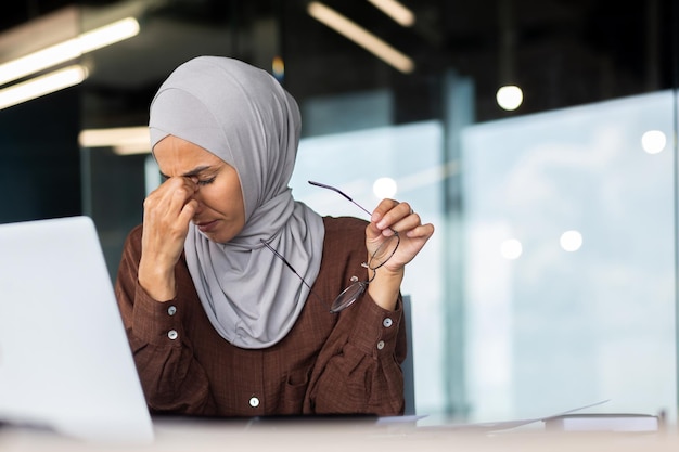 Une jeune enseignante musulmane fatiguée en hijab est assise au bureau devant un ordinateur portable, elle tient des lunettes en elle