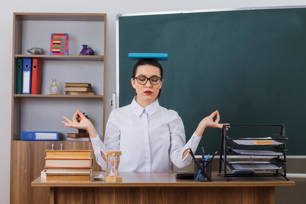 Jeune enseignante portant des lunettes assise au bureau de l'école devant le tableau noir dans la salle de classe avec un livre sur la tête essayant de se calmer en méditant