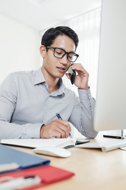 Jeune entrepreneur asiatique parlant au téléphone avec un partenaire commercial et écrivant dans un planificateur