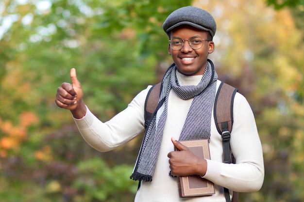 Jeune étudiant afro-américain africain noir, mec avec sac à dos et livre montrant le pouce vers le haut et souriant