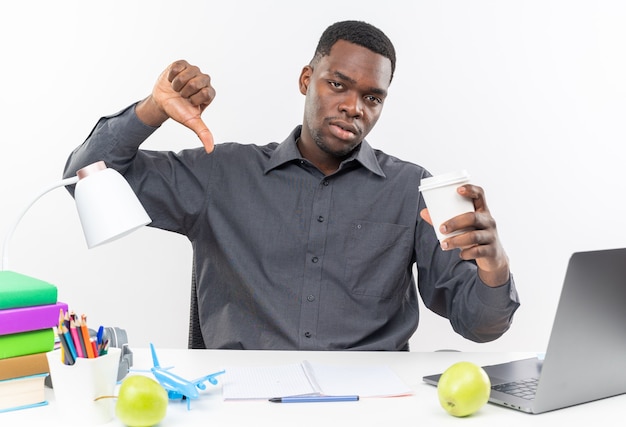 Jeune étudiant afro-américain bouleversé assis au bureau avec des outils scolaires tenant une tasse de papier et le pouce vers le bas