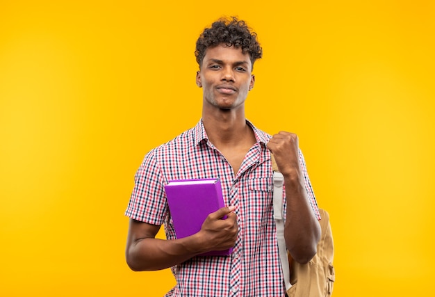 Jeune étudiant afro-américain confiant avec un sac à dos tenant un livre et gardant le poing