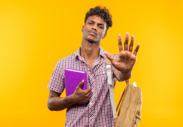 Jeune étudiant afro-américain confiant avec un sac à dos tenant un livre et un signe de la main d'arrêt gestuel isolé sur un mur orange avec espace de copie