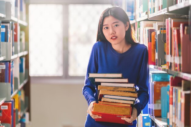 Jeune étudiant asiatique en costume décontracté tenant après avoir recherché le livre de l'étagère du livre dans la bibliothèque de l'université ou un collègue avec divers livres