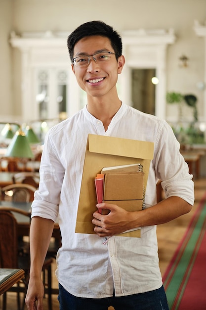 Jeune étudiant asiatique joyeux à lunettes avec des blocs-notes regardant joyeusement à huis clos dans la bibliothèque de l'université