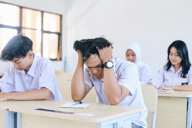 Jeune étudiant asiatique tenant la tête ayant des maux de tête alors qu'il était assis au bureau pendant l'examen à l'école