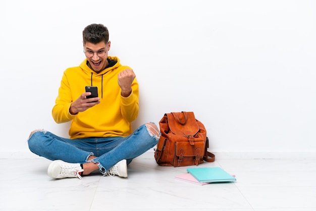 Jeune étudiant caucasien homme assis sur le sol isolé sur fond blanc surpris et envoyant un message