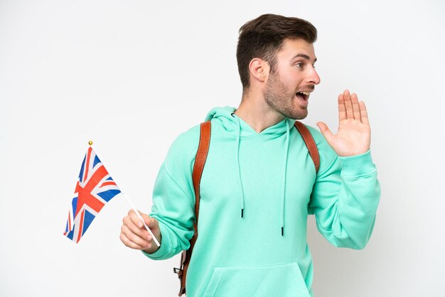 Jeune étudiant caucasien homme tenant un drapeau du Royaume-Uni isolé sur fond blanc criant avec la bouche grande ouverte sur le côté