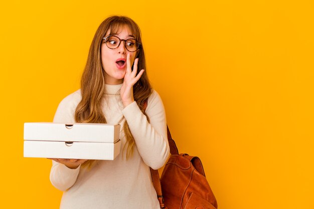 Jeune étudiant caucasien woman holding pizzas sur mur isolé dit un secret de freinage à chaud et à la recherche de côté