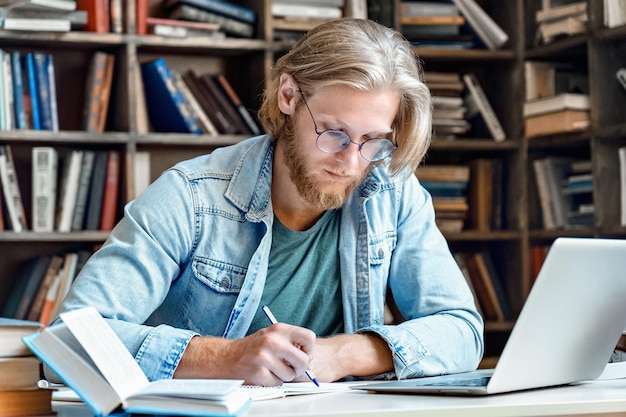 Jeune étudiant concentré au bureau de la bibliothèque, ordinateur portable moderne, écrire dans un cahier