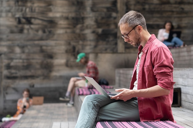Jeune étudiant concentré avec un ordinateur portable assis sur un banc en bois à l'extérieur de l'espace de coworking extérieur