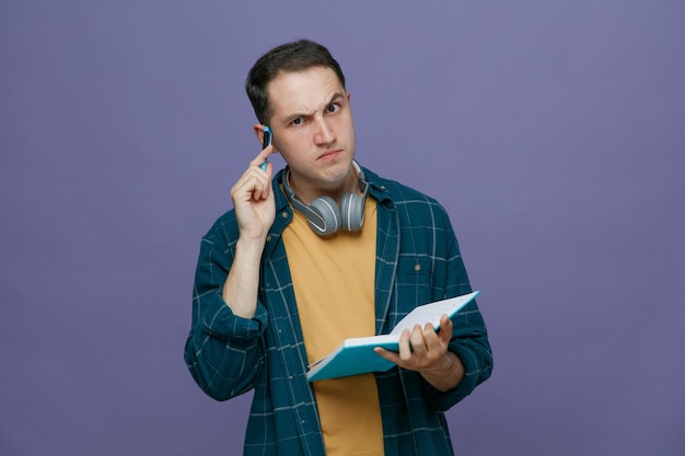 Jeune étudiant confus portant des écouteurs autour du cou tenant un carnet de notes regardant la caméra faisant un geste de réflexion avec un stylo isolé sur fond violet