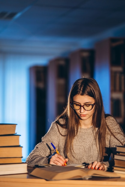 Jeune étudiant dans des verres se préparant à l'examen. Fille le soir est assise à une table dans la bibliothèque avec une pile de livres