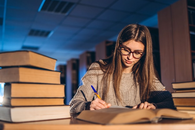 Jeune étudiant dans des verres se préparant à l'examen. Fille le soir est assise à une table dans la bibliothèque avec une pile de livres