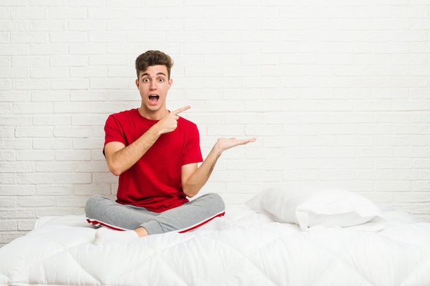 Jeune étudiant étudiant homme sur le lit excité tenant un espace de copie sur la paume.