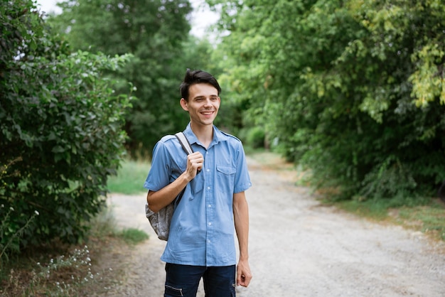 Jeune étudiant gai se tient dans une chemise bleue avec un sac à dos sur la route