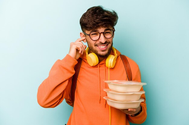 Jeune étudiant hispanique homme tenant un tupperware isolé sur fond bleu fond couvrant les oreilles avec les mains