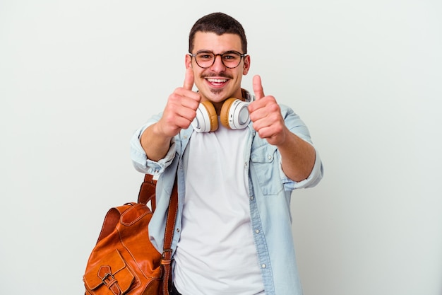 Jeune étudiant homme écoutant de la musique isolé sur un mur blanc avec des pouces vers le haut, des acclamations pour quelque chose