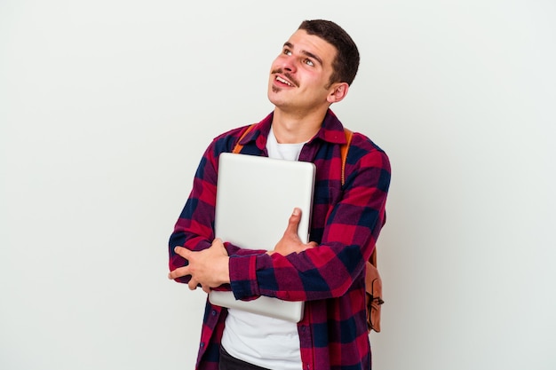 Jeune étudiant homme tenant un ordinateur portable isolé sur un mur blanc rêvant d'atteindre les objectifs et les fins