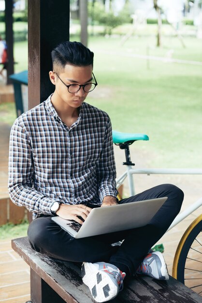 Jeune étudiant en informatique sérieux assis sur un banc sur le campus universitaire et travaillant sur un ordinateur portable