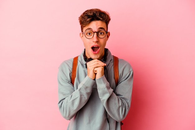 Jeune étudiant isolé sur fond rose priant pour la chance, étonné et ouvrant la bouche regardant vers l'avant.