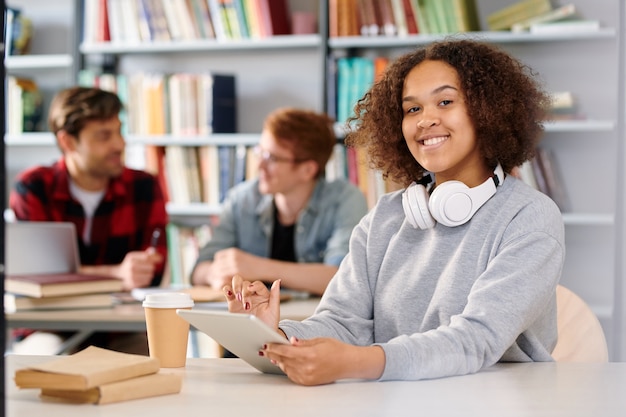 Jeune étudiant joyeux avec tablette se prépare pour l'examen tout en travaillant dans la bibliothèque du collège