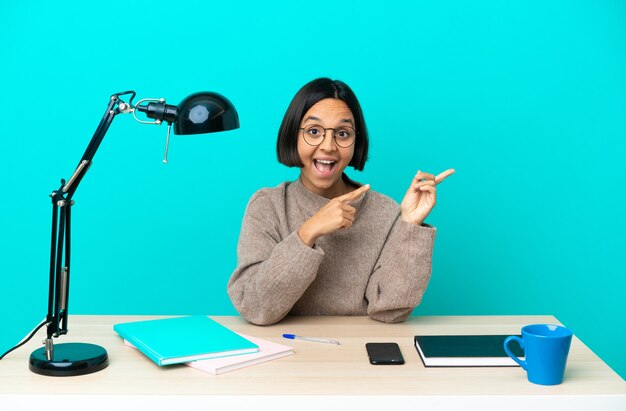 Jeune étudiant métisse femme étudiant sur une table effrayée et pointant vers le côté