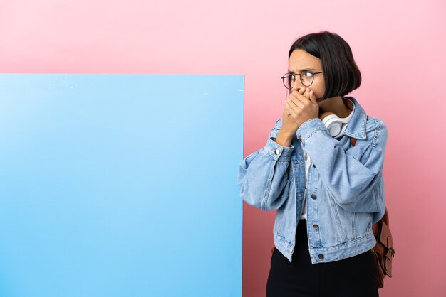 Jeune étudiant métisse femme avec une grande bannière sur mur isolé couvrant la bouche et à la recherche sur le côté